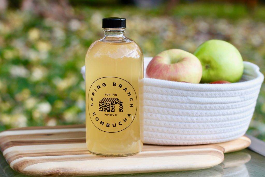 Photo of 16 oz. glass growler filled with Apple Pie kombucha, with a white basket of various apples in the background.