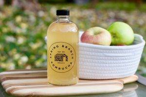 Photo of 16 oz. glass growler filled with Apple Pie kombucha, with a white basket of various apples in the background.